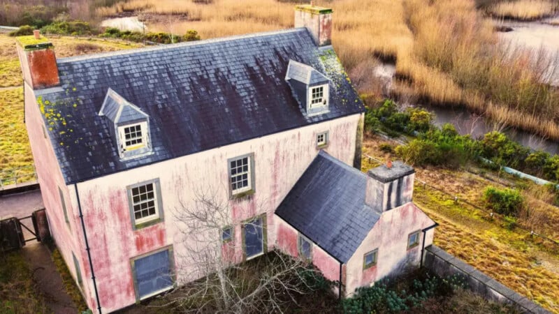 A large, old pink house with a slate roof and dormer windows, showing signs of wear, sits beside a marshy, grassy field with water channels in the background. A leafless tree stands in front of the house.