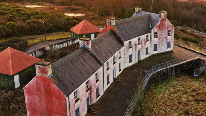 An aerial view of an abandoned, weathered building with faded red and white paint, boarded-up windows, and moss on the roof, surrounded by overgrown grass and bushes in a rural landscape.