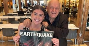 A smiling young woman holds a vintage "EARTHQUAKE" movie poster while an older man stands behind her with his hand on her shoulder in a busy cafeteria or food court setting.