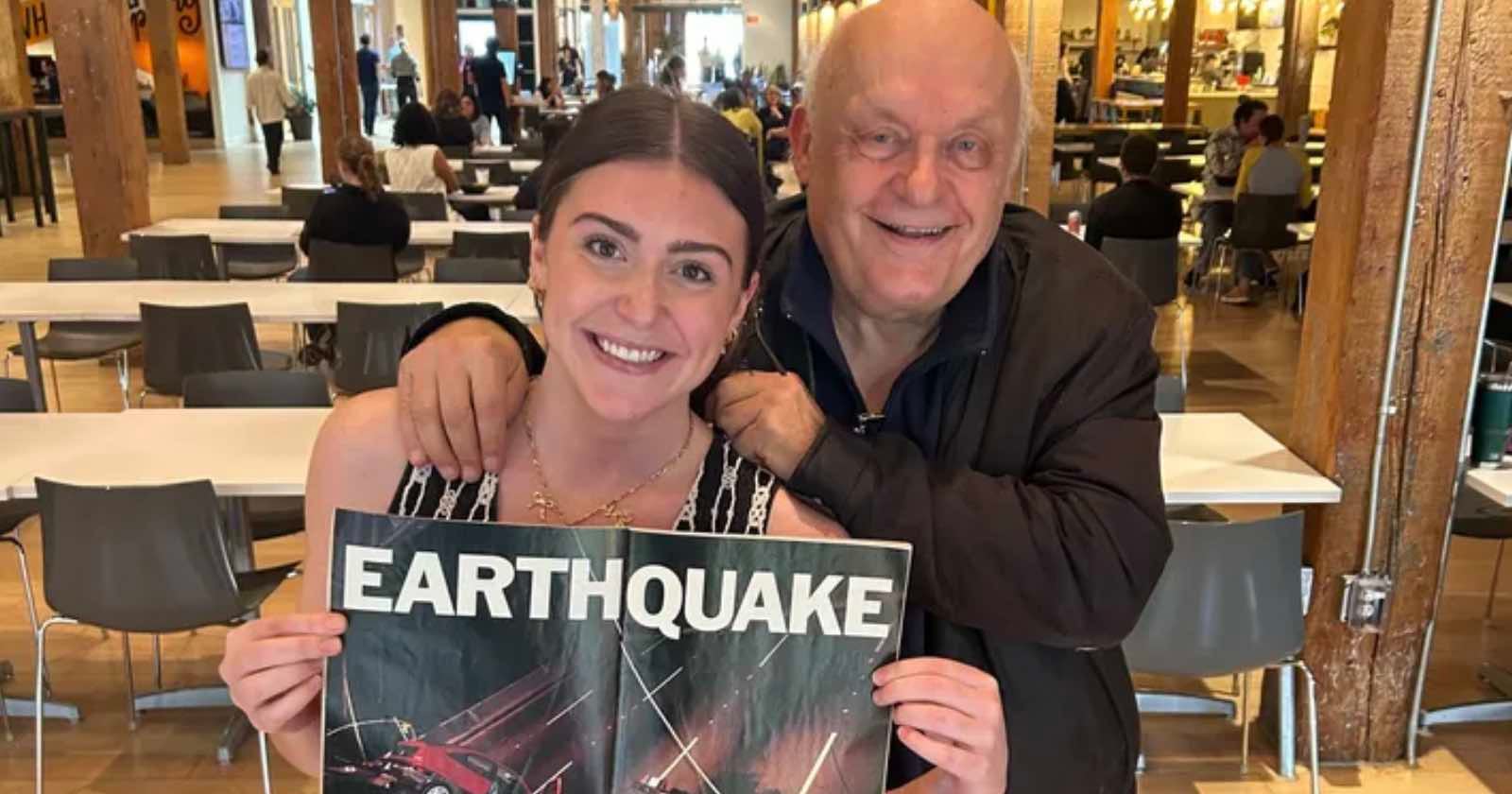 A smiling young woman holds a vintage "EARTHQUAKE" movie poster while an older man stands behind her with his hand on her shoulder in a busy cafeteria or food court setting.
