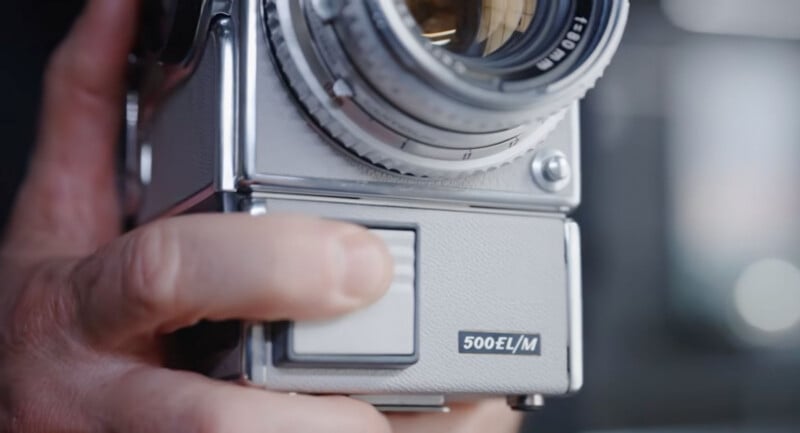 A close-up of a hand holding a vintage silver Hasselblad 500EL/M camera, focusing on the camera's front controls and textured grip.