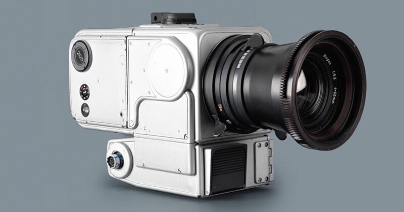 A large, boxy silver camera with a prominent lens and several buttons, dials, and screws on its side, set against a plain gray background.