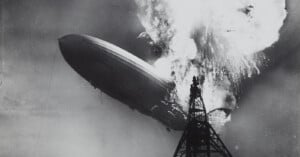 A large airship bursts into flames near a tall mooring mast, with thick smoke and fire engulfing its upper section against the sky in this dramatic black-and-white photo.