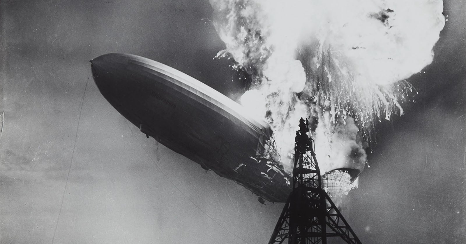 A large airship bursts into flames near a tall mooring mast, with thick smoke and fire engulfing its upper section against the sky in this dramatic black-and-white photo.