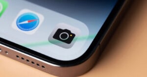Close-up of a smartphone screen showing the Camera app icon, Safari browser icon, and part of another app, with the phone’s edge and speaker holes visible on a light-colored surface.