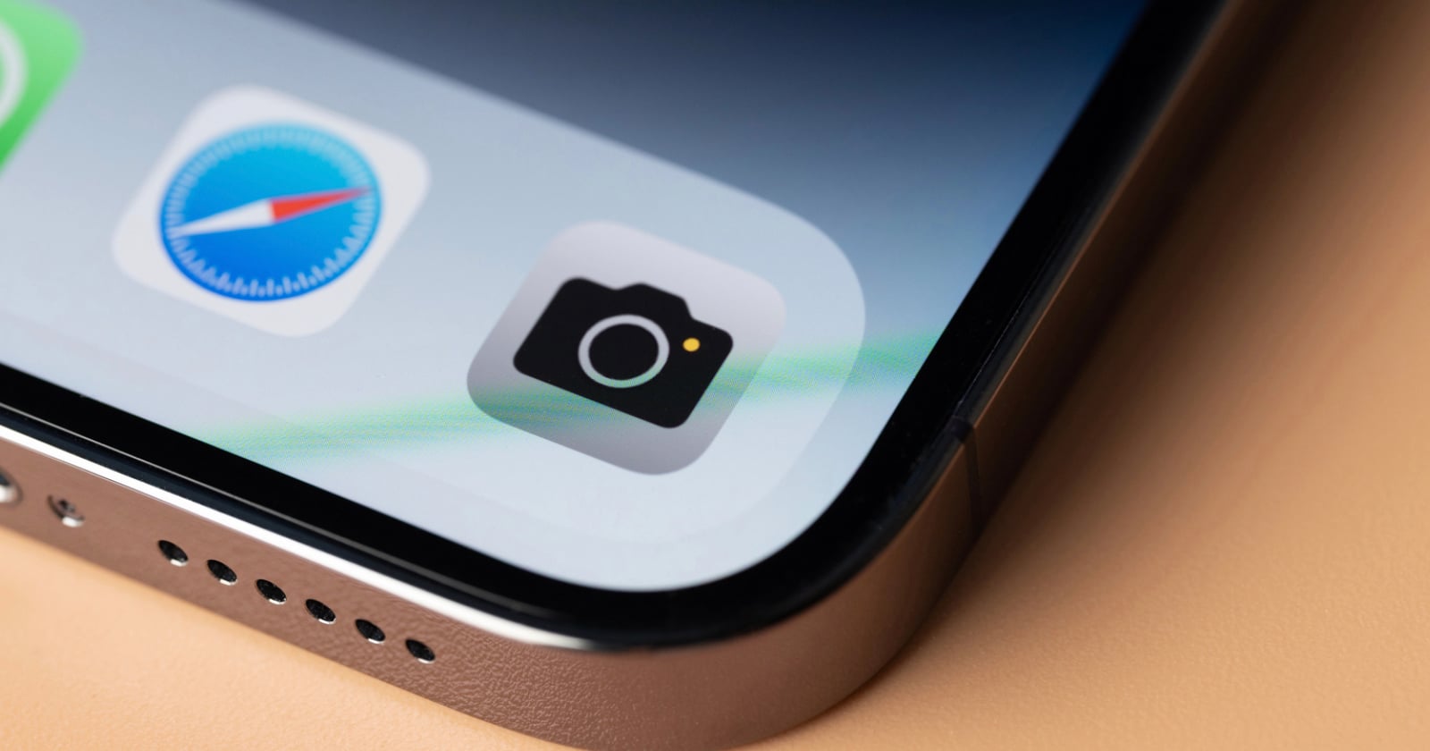 Close-up of a smartphone screen showing the Camera app icon, Safari browser icon, and part of another app, with the phone’s edge and speaker holes visible on a light-colored surface.
