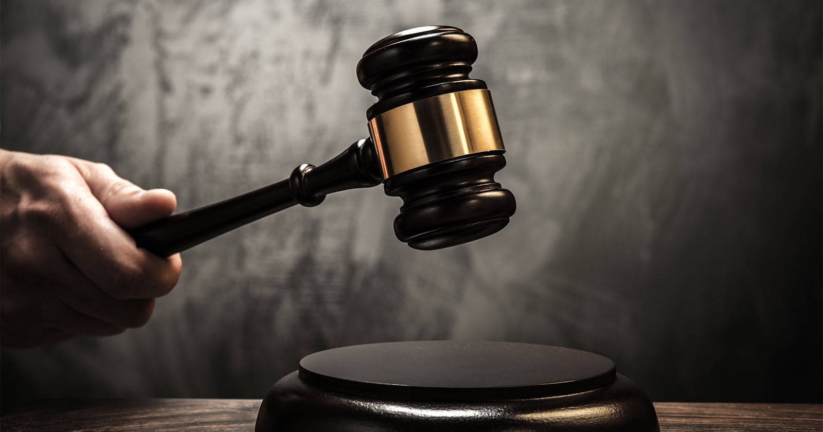 A hand holding a wooden judge’s gavel above a sound block, set against a dark, blurred background, symbolizing law and justice.