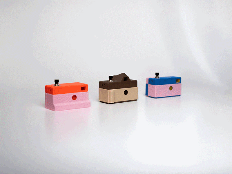 Three small, colorful toy cameras with simple boxy shapes are displayed on a white surface. The cameras are pink and orange, beige and brown, and pink and blue. The background is plain white.