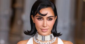 A woman with shoulder-length dark hair styled in soft waves wears a pearl choker and a white outfit, posing against a blurred background. Her makeup is glamorous with defined brows and nude lips.