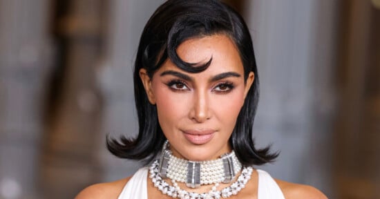 A woman with shoulder-length dark hair styled in soft waves wears a pearl choker and a white outfit, posing against a blurred background. Her makeup is glamorous with defined brows and nude lips.