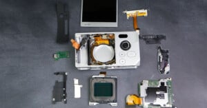 A disassembled digital camera with its various components, including the lens assembly, circuit boards, buttons, and LCD screen, laid out neatly on a dark surface.