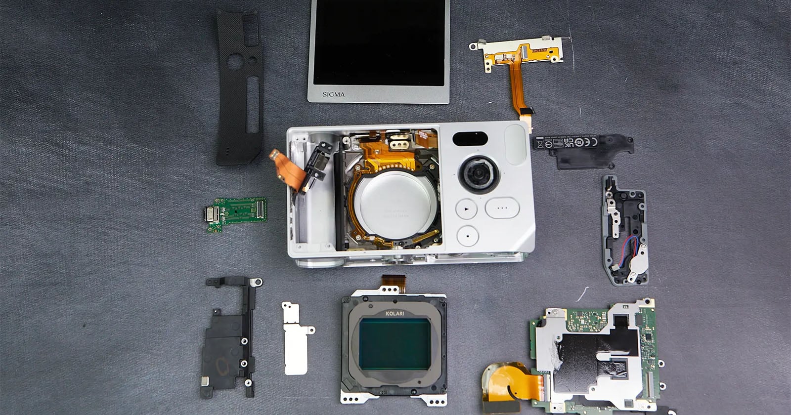 A disassembled digital camera with its various components, including the lens assembly, circuit boards, buttons, and LCD screen, laid out neatly on a dark surface.