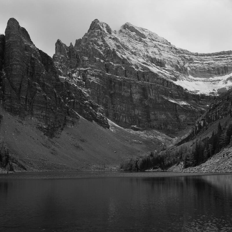 A dramatic black-and-white photo of a rugged mountain with steep cliffs and patches of snow, reflected in the calm waters of a lake below, surrounded by sparse trees.