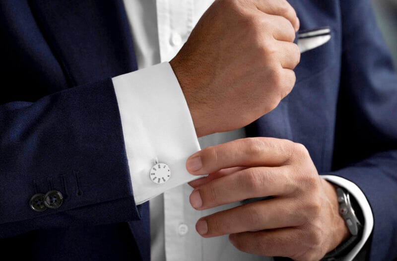 A person in a navy blue suit adjusts their white shirt cuff, which has a white round cufflink. A white pocket square is visible in the suit jacket's breast pocket.
