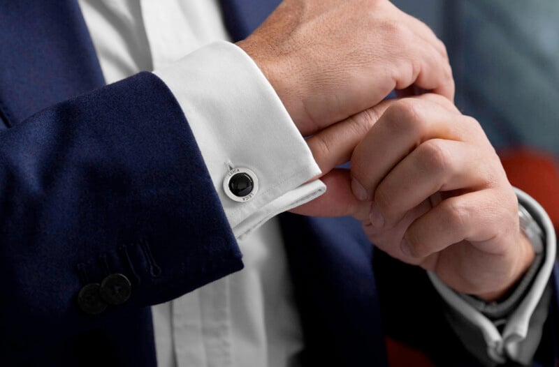 A person in a dark blue suit jacket adjusts a white dress shirt cuff with a silver and black cufflink. The focus is on the hand and sleeve, suggesting attention to detail and formal attire.