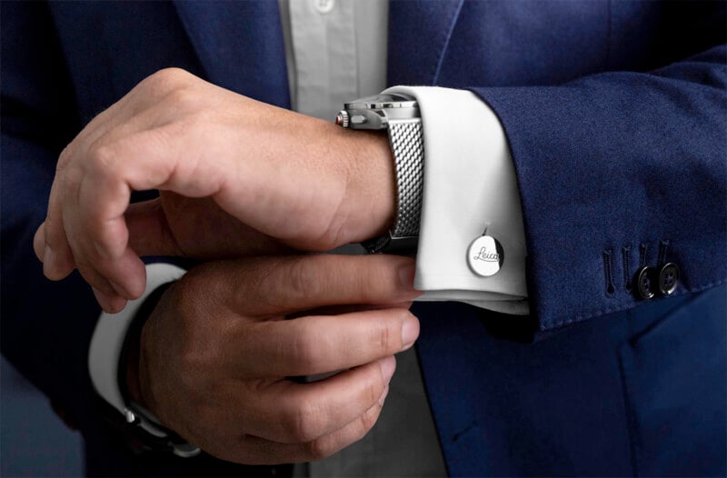 A person in a blue suit jacket adjusts a silver mesh watch on their wrist, with a white shirt cuff showing a round cufflink.