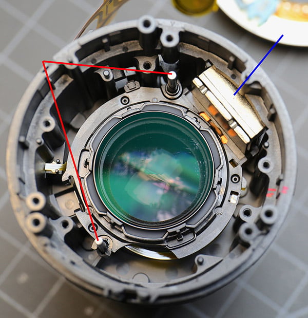 Close-up of a disassembled camera lens showing internal components, with red and blue lines drawn to highlight specific parts and pathways inside the lens housing.