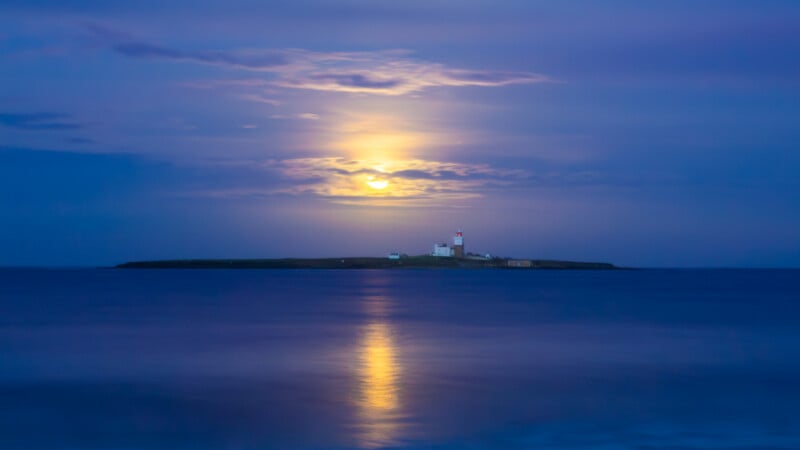 A small island with a lighthouse is silhouetted against a twilight sky. The moon glows above, casting a bright reflection on the calm, blue water surrounding the island.