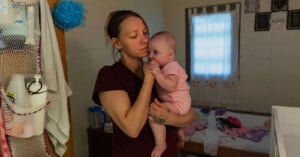 A woman in a maroon shirt holds a baby dressed in pink in a modest, dimly lit bedroom. The woman looks thoughtful as the baby puts her hand in her mouth. The bed behind them is unmade, with baby items scattered around.