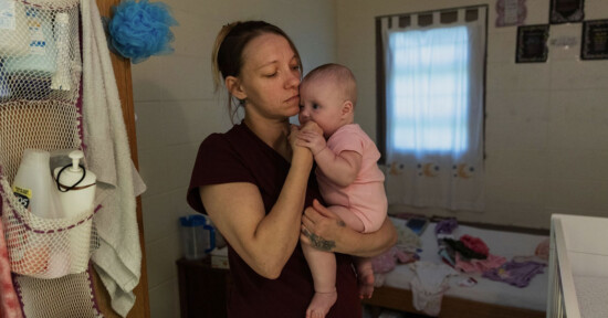 A woman in a maroon shirt holds a baby dressed in pink in a modest, dimly lit bedroom. The woman looks thoughtful as the baby puts her hand in her mouth. The bed behind them is unmade, with baby items scattered around.