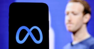 A smartphone displays the Meta logo in the foreground, with a blurred person standing against a blue background in the distance.