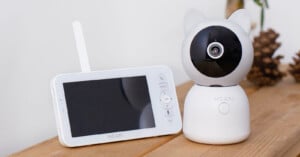A white baby monitor with a screen and a camera shaped like a cat, both labeled “MEARI,” are placed on a wooden surface next to some pinecones.