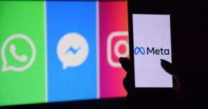 A hand holds a smartphone displaying the Meta logo, with blurred logos of WhatsApp, Messenger, and Instagram shown on a colorful screen in the background.