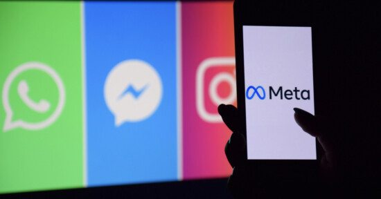A hand holds a smartphone displaying the Meta logo, with blurred logos of WhatsApp, Messenger, and Instagram shown on a colorful screen in the background.