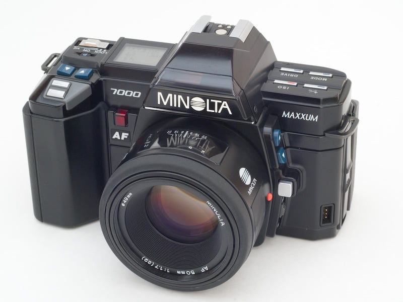 A black Minolta Maxxum 7000 autofocus film camera with a 50mm lens, featuring various buttons and dials, displayed on a plain white background.