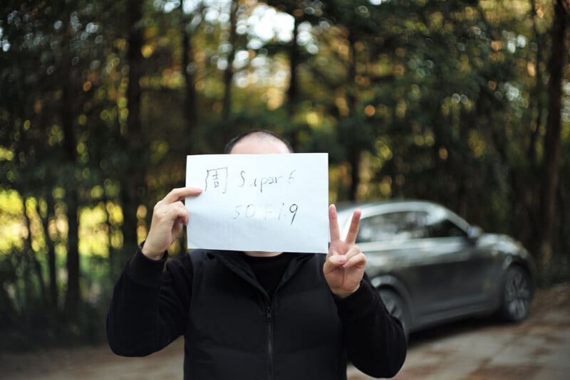 A person standing outdoors holds a handwritten sign in front of their face, with one hand making a peace sign. A silver car and trees are visible in the blurred background.