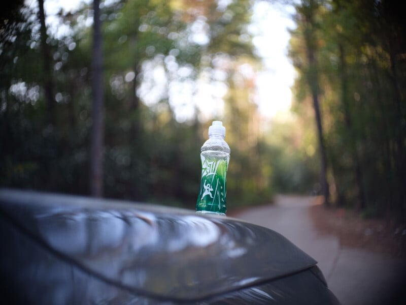 A plastic bottle filled with green liquid sits on the roof of a car, with a blurred forest and pathway in the background.