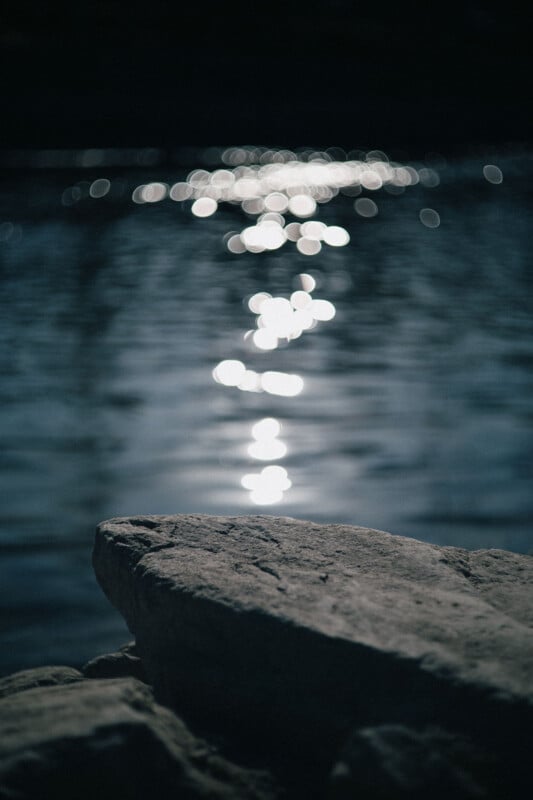 A close-up of a rock in the foreground with soft focus, while sunlight reflects on rippling water in the background, creating bright, blurry light spots. The overall mood is calm and serene.