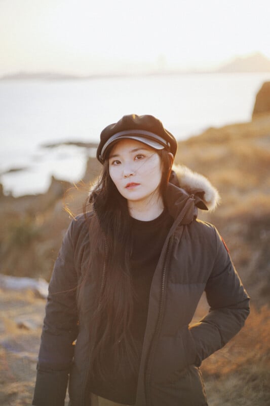 A woman with long dark hair wearing a black winter coat with a fur-trimmed hood and a black cap stands outdoors near water and rocky terrain at sunset.
