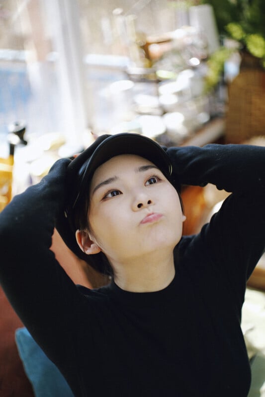 A young person wearing a black shirt and cap sits indoors with hands resting on their head, cheeks puffed out and lips pursed in a playful or thoughtful expression. The background is softly blurred.