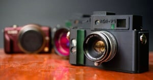 Three vintage cameras are displayed on a wooden surface, with one black camera in sharp focus in the foreground and two blurred cameras in the background against a dark backdrop.