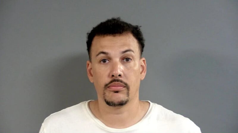 A man with short dark hair, a goatee, and mustache, wearing a plain white t-shirt, stands against a plain gray background looking directly at the camera.