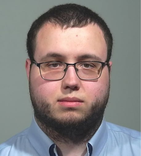 A young man with short brown hair, a beard, and glasses is wearing a light blue collared shirt, looking directly at the camera with a neutral expression against a plain background.
