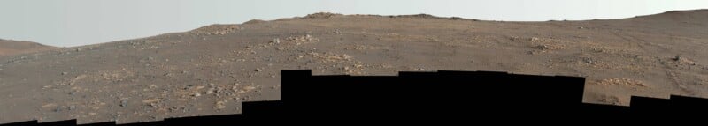 A rocky, reddish-brown Martian landscape with scattered stones and gentle slopes under a pale sky; the image has a black, uneven section along the bottom edge.