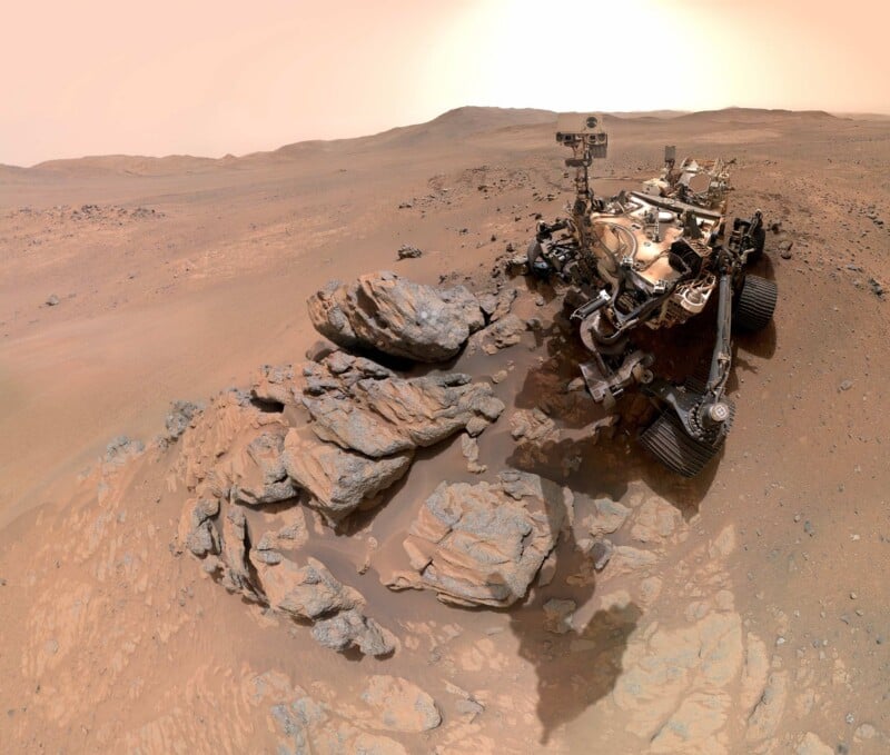 A Mars rover stands beside scattered rocks on the dusty, reddish surface of Mars under a hazy sky, with barren hills visible in the background.