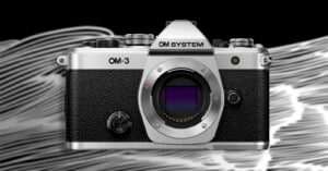 A silver and black OM System OM-3 camera is shown facing forward against a background of abstract white and black swirling lines. The camera lens mount is visible without a lens attached.