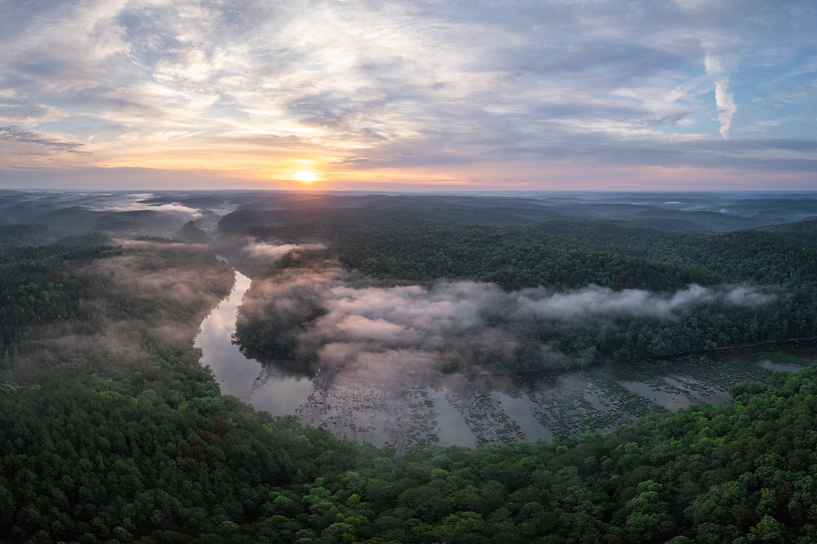 Aerial view of a lush green forest with a winding river, mist rising above the water, and the sun rising on the horizon under a partly cloudy sky.
