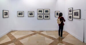 A person with a light brown backpack stands in a white-walled gallery, looking at framed black-and-white photographs displayed on the walls. The floor has a geometric tile pattern.