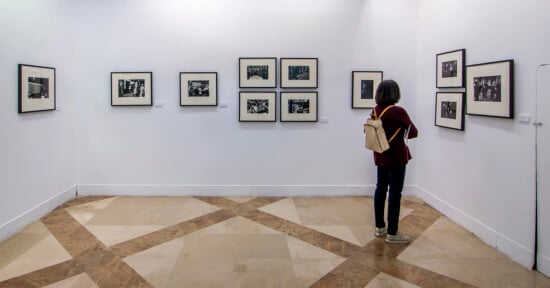 A person with a light brown backpack stands in a white-walled gallery, looking at framed black-and-white photographs displayed on the walls. The floor has a geometric tile pattern.