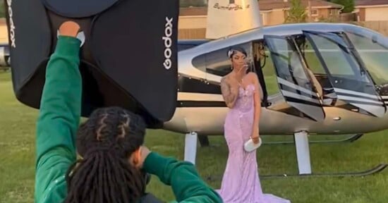 A young woman in a light purple gown poses for a photo in front of a helicopter, holding a small clutch. A photographer with a flash equipment is capturing the moment outdoors.