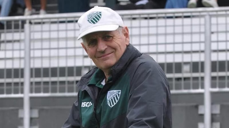 An older man in a white cap and dark jacket with matching green and white sports logos smiles outdoors in front of metal railings and bleachers.