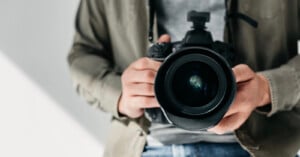 A person wearing a khaki jacket and gray shirt is holding a professional camera with both hands, pointing the lens directly toward the viewer.