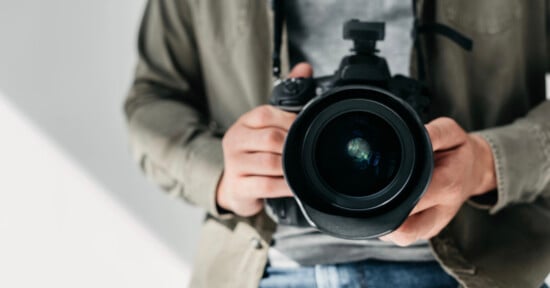 A person wearing a khaki jacket and gray shirt is holding a professional camera with both hands, pointing the lens directly toward the viewer.