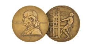 Two sides of a gold Pulitzer Prize medal: one shows Benjamin Franklin’s portrait with "Honoris Causa," the other depicts a man at a printing press with engraved text about American journalism.