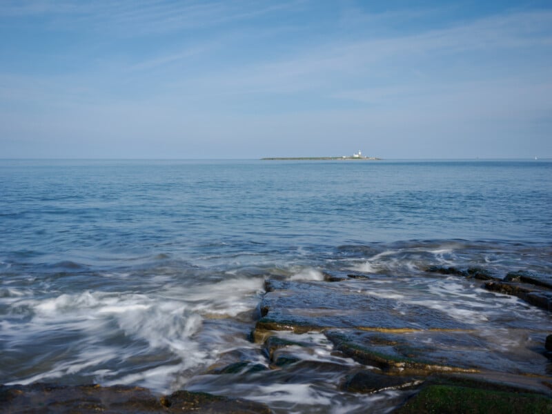 Waves gently wash over rocky shore in the foreground, with a calm blue sea stretching to the horizon. In the distance, a small island with a lighthouse sits under a partly cloudy sky.