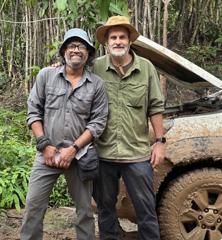 Two men in outdoor clothing and hats stand next to a muddy vehicle with the hood up, surrounded by dense green foliage in a jungle setting. Both are smiling and appear relaxed.